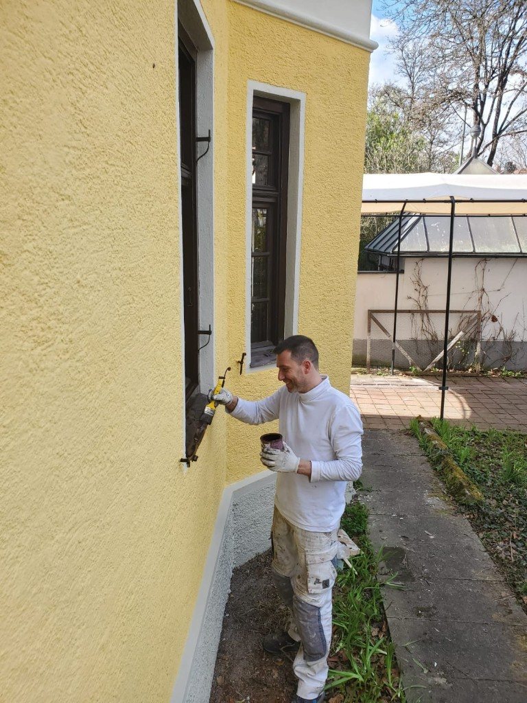 Florian Hemetzberger bei Lackierarbeiten an einem Fenstergitter vor einer gelben Fassade